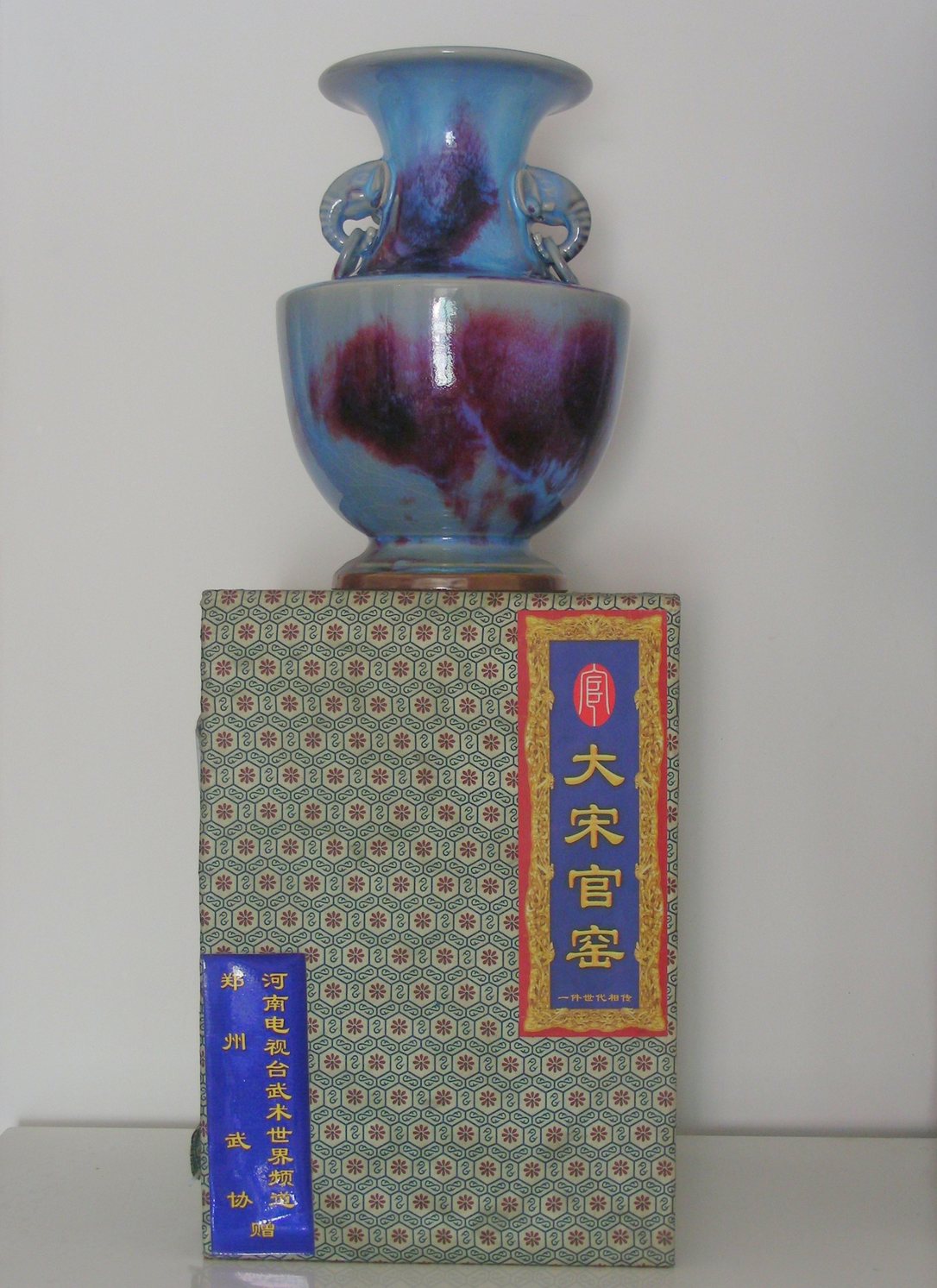 「大宋官窯」と書かれた箱の上に載せた、水色に紫が流れる釉薬の丸い壺